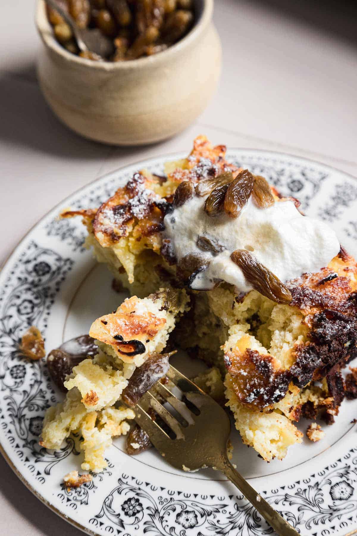 A piece of noodle kugel topped with whipped cream and raisins is on an ornate white plate. A fork rests on top, cutting into the dessert. A small bowl of raisins is blurred in the background.