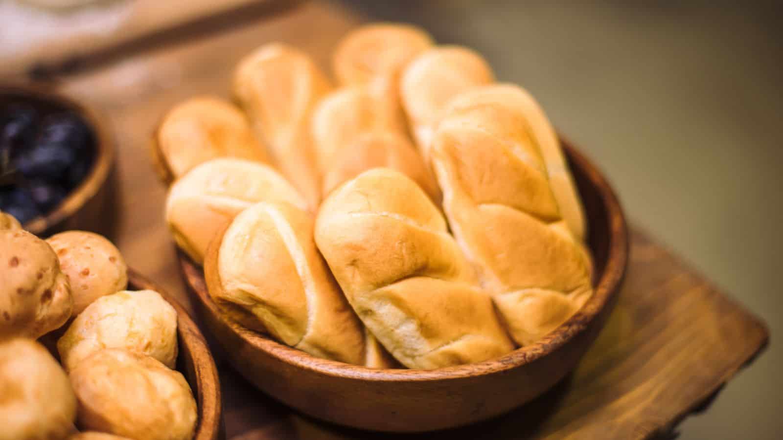 A wooden bowl filled with freshly baked bread rolls is placed on a wooden surface. Another bowl with round buns is partially visible on the left. The bread rolls have a golden-brown crust. The background is softly blurred.