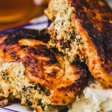 Two pieces of browned, stuffed chicken breast sit on a plate next to a serving of mashed potatoes. The stuffing appears to contain herbs and possibly cheese.