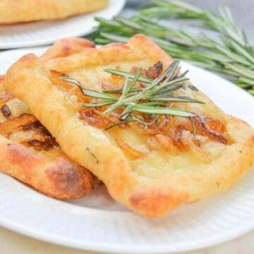 Rectangular appetizers topped with caramelized onions and garnished with rosemary sit on a white plate. A sprig of fresh rosemary lies beside the plate on a light-colored surface. Another plate with pastries is partially visible in the background, sure to bring a smile even to your mother-in-law.