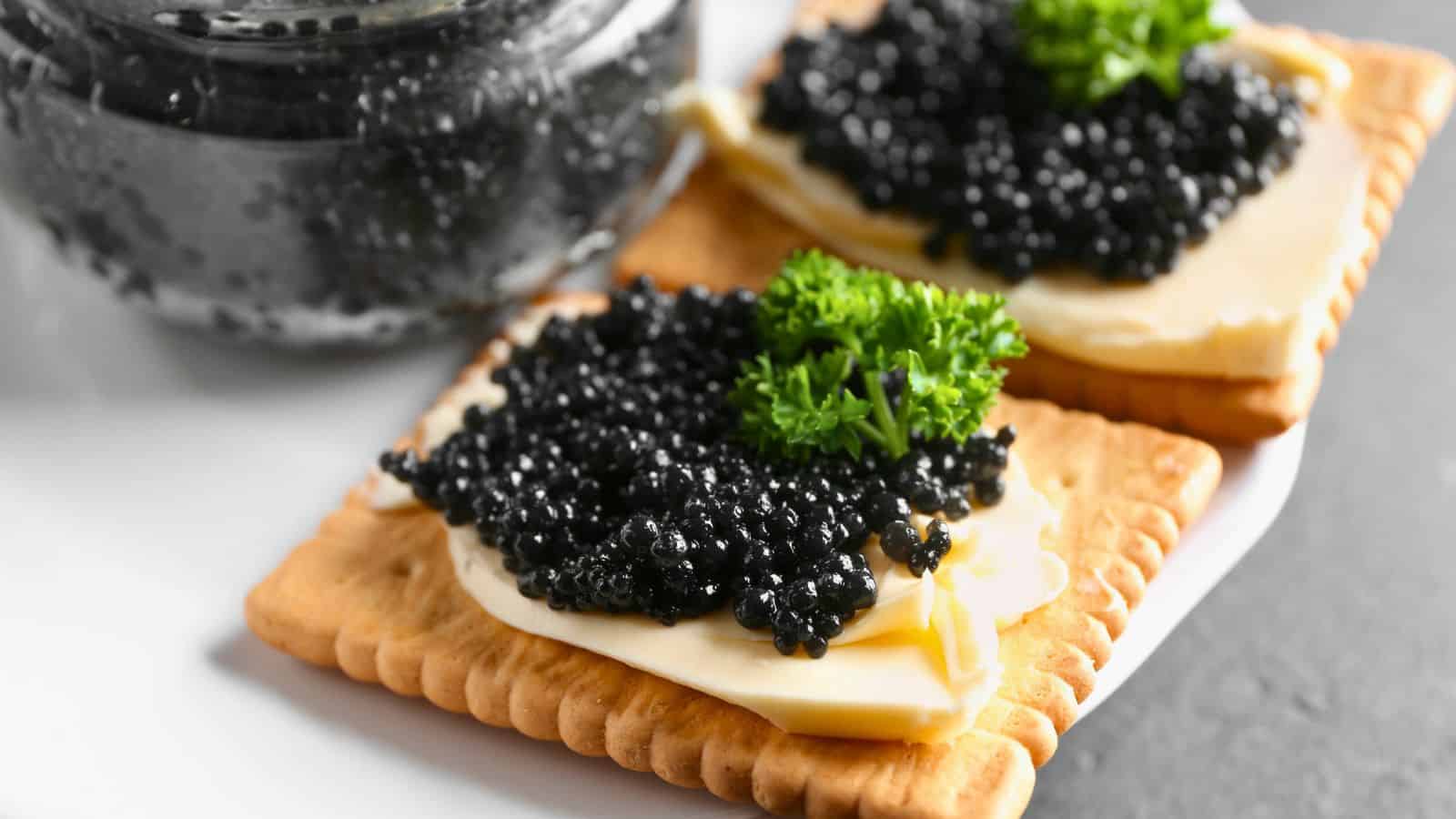 Two square crackers topped with slices of cheese, black caviar, and garnished with parsley are on a white plate. A jar of black caviar is partially in view beside the plate.