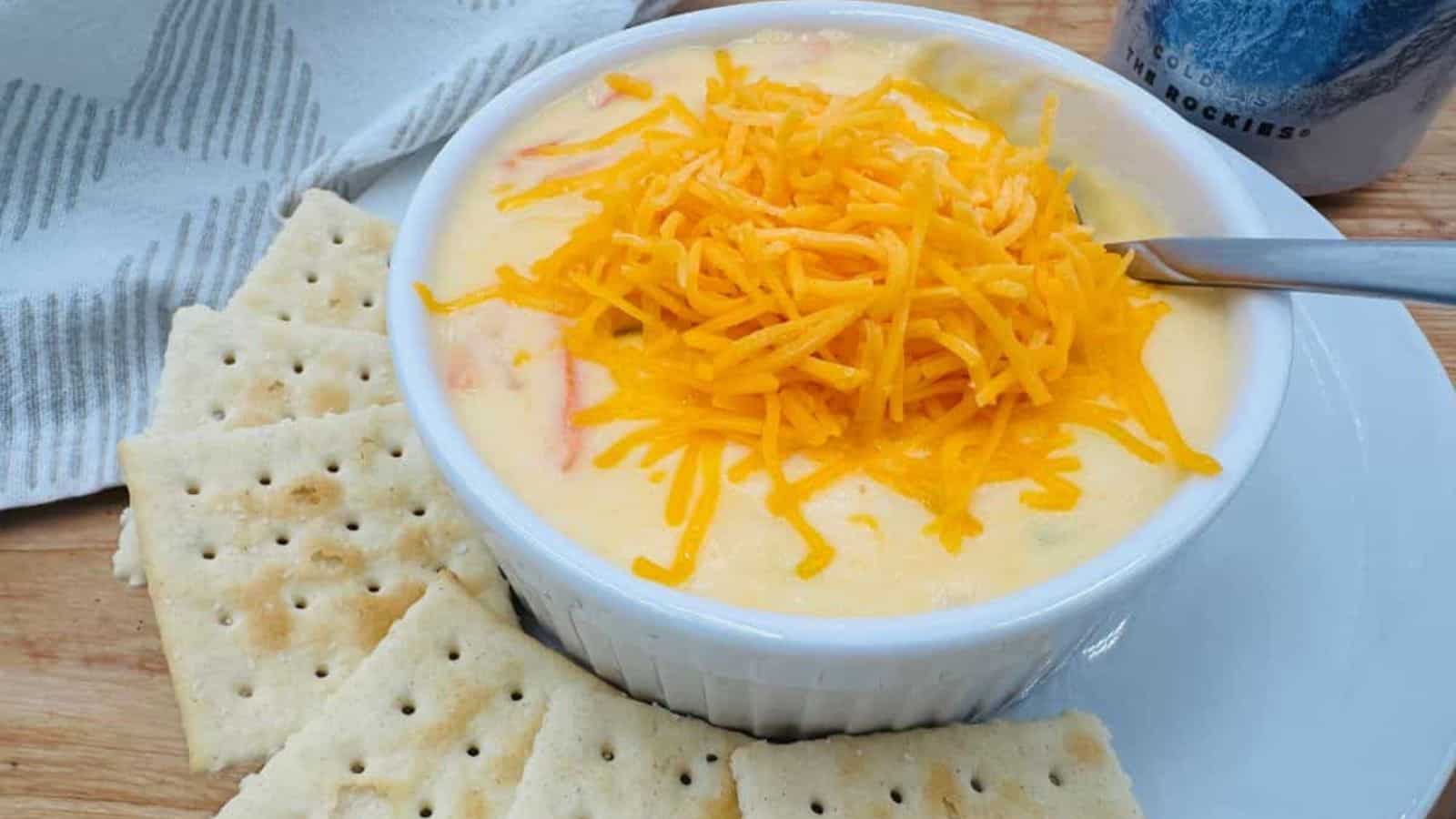 A white ramekin filled with creamy soup or dip, topped with shredded cheddar cheese, is surrounded by saltine crackers on a white plate. Perfect for family meals, a spoon rests in the ramekin and a striped napkin is nearby.