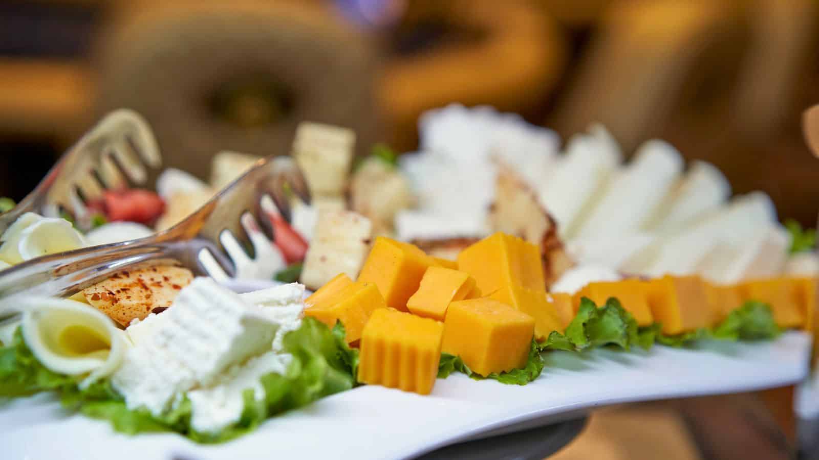 A close-up of a cheese platter featuring various types of cheese, including white, yellow, and orange cubes and slices. The arrangement is garnished with lettuce leaves and accompanied by serving tongs.