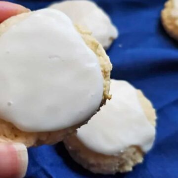 A hand holds a round homemade cookie with white icing. Several similar cookies with white icing rest on a dark blue cloth in the background, reminiscent of classic childhood cookies.