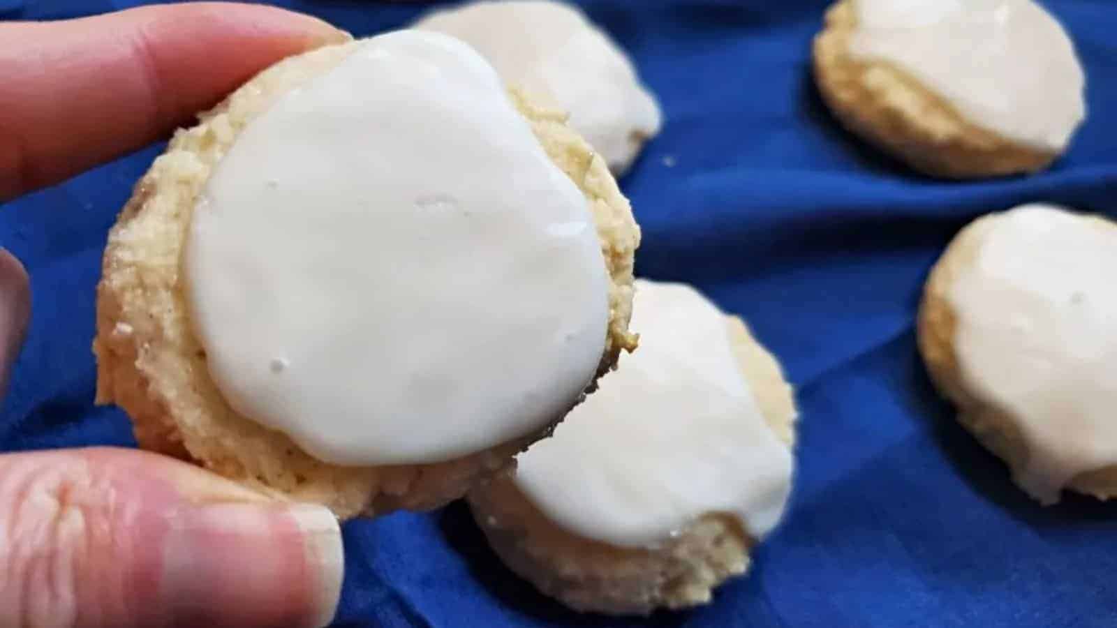 A hand holds a round homemade cookie with white icing. Several similar cookies with white icing rest on a dark blue cloth in the background, reminiscent of classic childhood cookies.