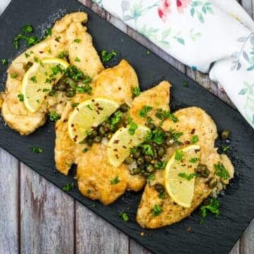 Grilled chicken fillets topped with lemon slices and herbs are arranged on a black slate platter. The platter is on a wooden surface with a floral-patterned cloth partially visible on the side.