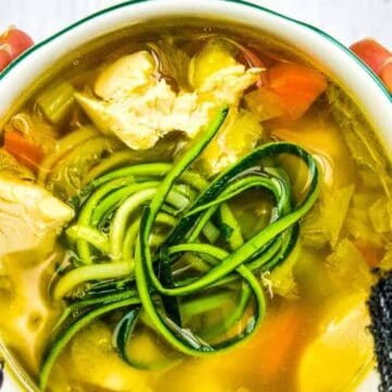A pair of hands holding a bowl of clear, low-carb soup. The soup features pieces of chicken, sliced celery, carrots, and spiraled zucchini. The person is wearing a dark knitted sweater against a background of white wooden dishes.