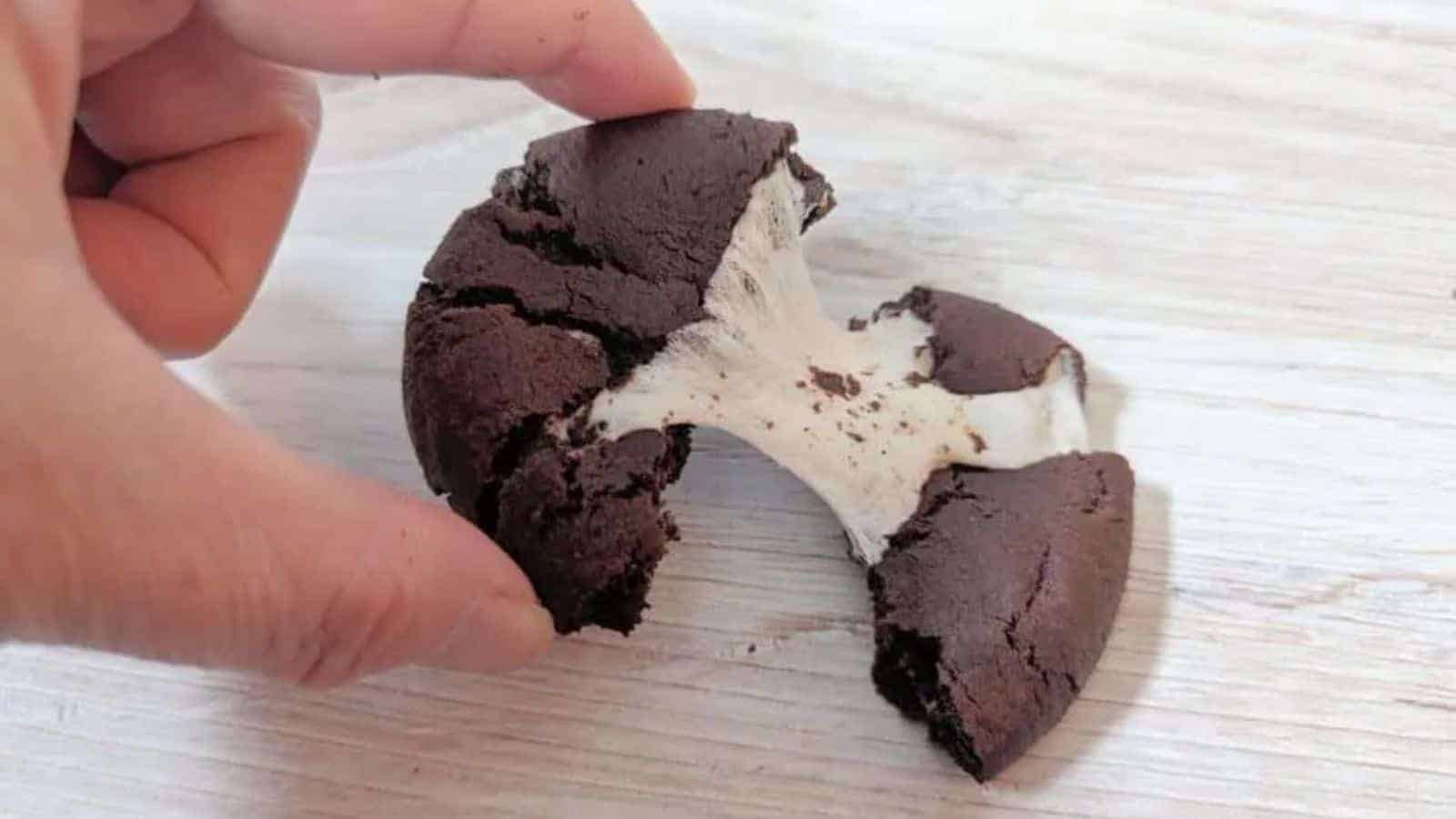 A hand pulls apart a chocolate homemade cookie, revealing a stretchy marshmallow filling inside. The cookie is on a light wooden surface, bringing back memories of classic childhood cookies.