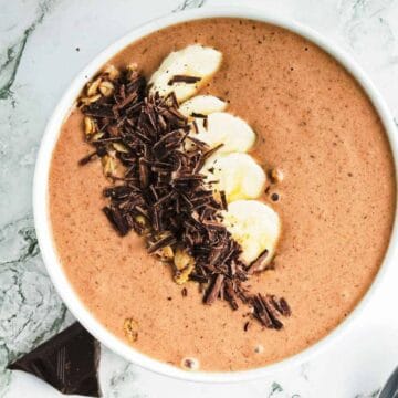 A bowl filled with a chocolate smoothie, topped with banana slices, granola, and chocolate shavings, sits on a white marble surface next to a spoon and a piece of chocolate.