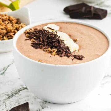 A white bowl filled with a chocolate smoothie, topped with banana slices, granola, and chocolate shavings sits on a marble surface. A bowl of granola and pieces of chocolate are nearby.