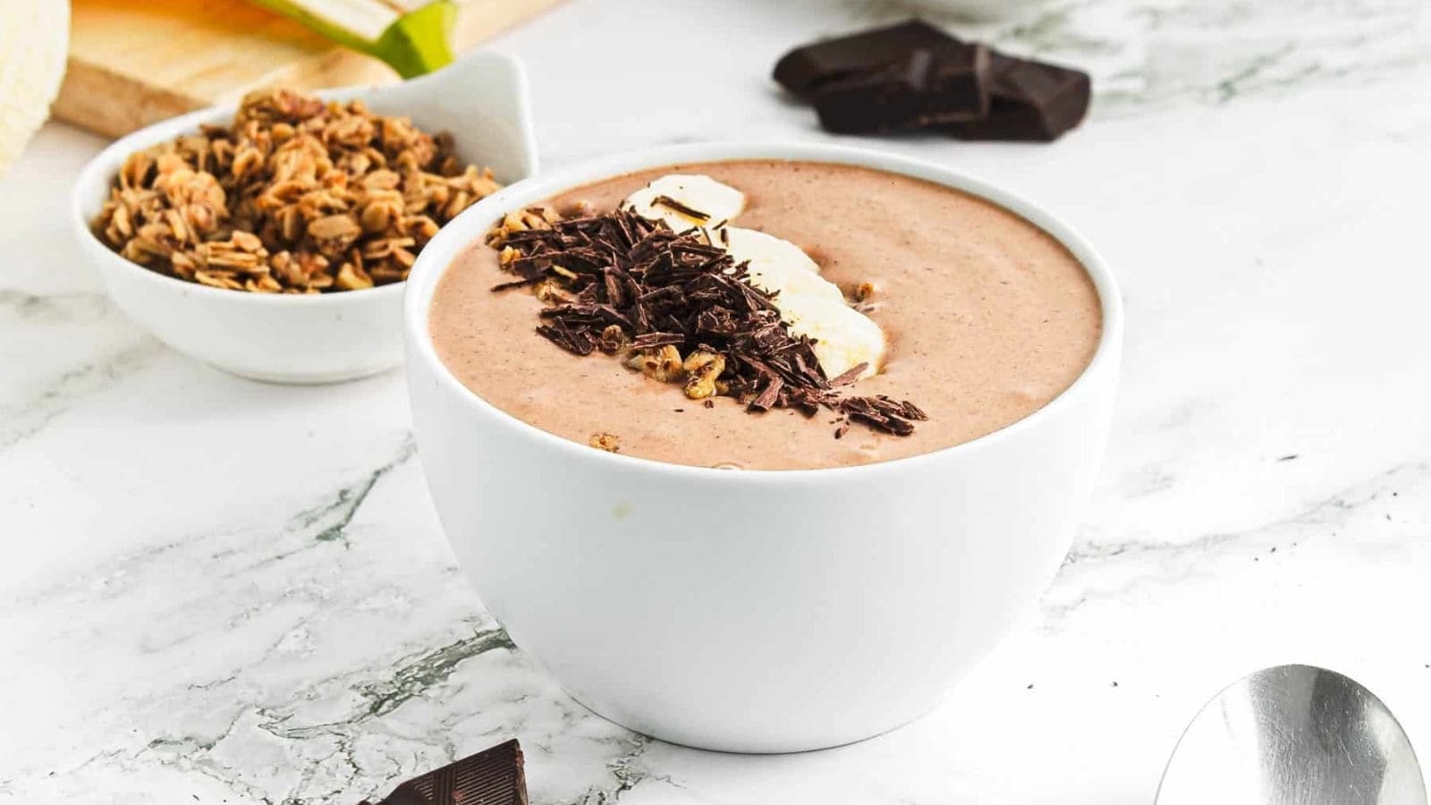 A bowl filled with a chocolate smoothie, topped with banana slices, granola, and chocolate shavings, sits on a white marble surface next to a spoon and a piece of chocolate.