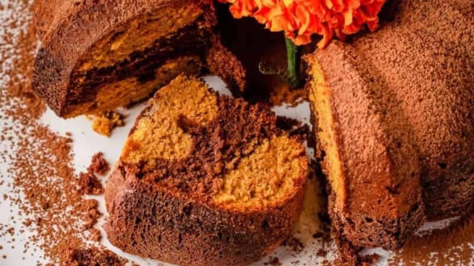 A sliced marble bundt cake with a swirl of chocolate and vanilla layers is topped with cocoa powder, decorated with an orange marigold flower—perfect for Mother’s Day cakes. Cake crumbs and cocoa powder are scattered on the surface.