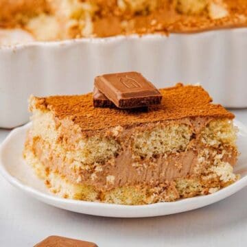 A slice of chocolate tiramisu sits on a white plate, topped with two pieces of chocolate. The dessert has visible layers of cream and cake. The background shows a baking dish and chocolate pieces on the table.