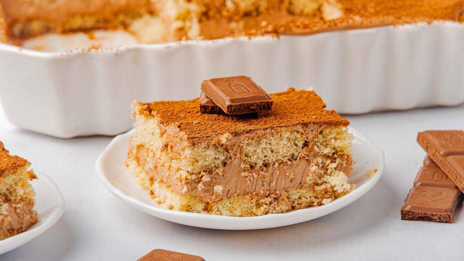 A slice of layered tiramisu with cocoa powder and a chocolate square on top, served on a white plate. A baking dish with more tiramisu is in the background.