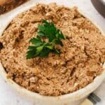 A bowl filled with classic Jewish chopped chicken livers, garnished with a fresh sprig of parsley. The texture appears crumbly, with visible small chunks. A knife with a light-colored handle is partially visible next to the bowl.