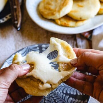 A person pulls apart a small, round Colombian arepa filled with melted cheese, showing the cheese stretching between the two halves. More Colombian arepas sit on a white plate in the background.
