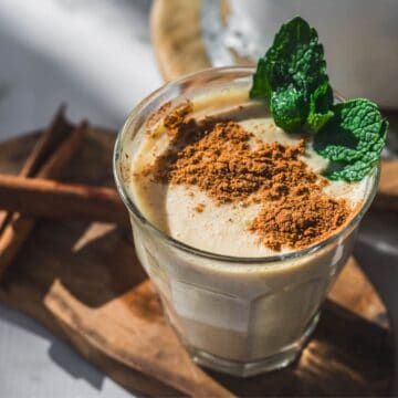 A glass of creamy Colombian Avena, a traditional Colombian drink, topped with ground cinnamon and garnished with fresh mint leaves, sits on a wooden surface next to two cinnamon sticks.