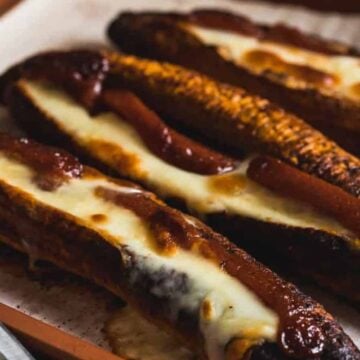 Three baked bananas on a parchment-lined tray, split open and filled with melted cheese and a dark sauce, showing caramelized and slightly charred edges.