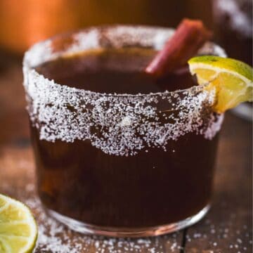 A glass with a salted rim filled with a dark Canelazo Cocktail, garnished with a cinnamon stick and a lime wedge. A lime slice sits next to the glass on a wooden surface, showcasing this classic Colombian Cocktail.