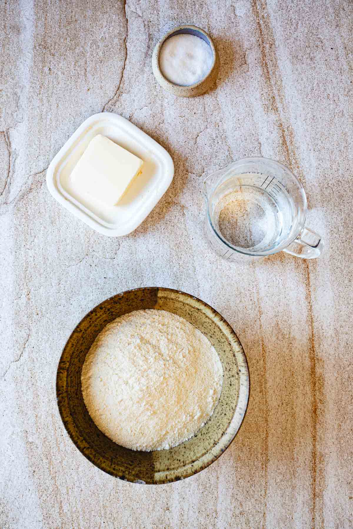 A bowl of flour, a small dish of butter, a cup of water, and a small bowl of salt are arranged on a light-colored, textured surface—essential ingredients for making Colombian arepas, a beloved traditional Colombian food.