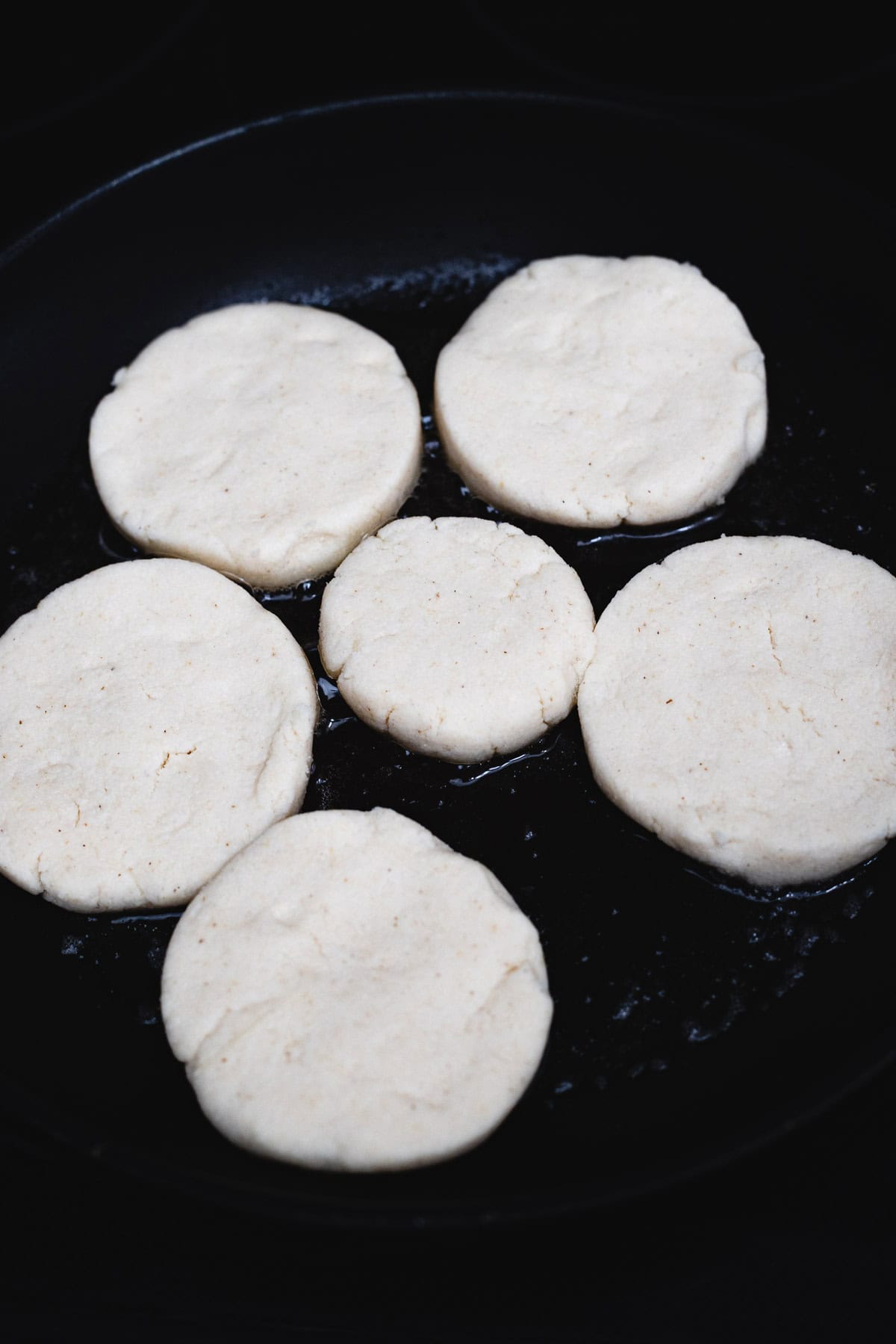 Six uncooked, round pieces of dough are arranged in a black skillet, spaced apart and ready to be cooked. The smooth, pale rounds are perfect for trying an authentic Colombian Arepas recipe and enjoying classic Colombian food.