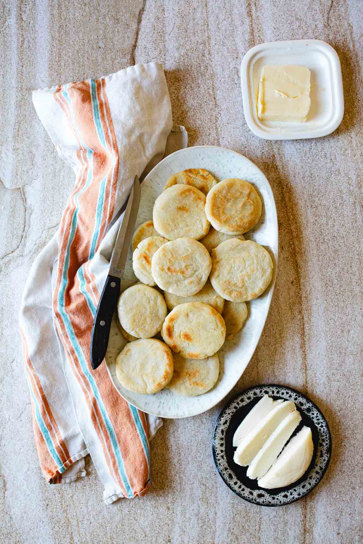 A plate of round, golden-brown Colombian arepas sits beside a butter knife and a striped cloth; a small dish with butter pats and a separate plate with sliced cheese are also on the table.