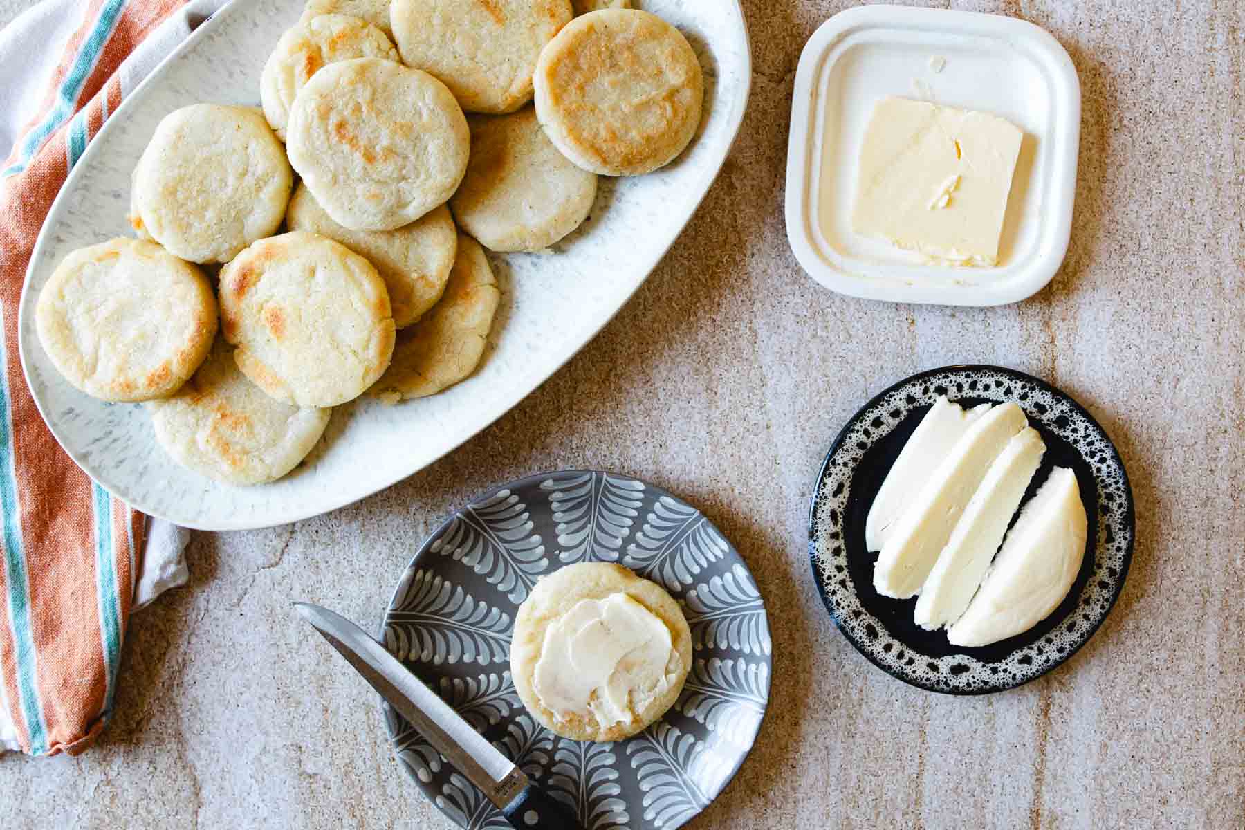 A plate of round, golden Colombian arepas next to a striped towel, a dish of butter with a butter knife, and a plate holding slices of white cheese on a light-colored surface.