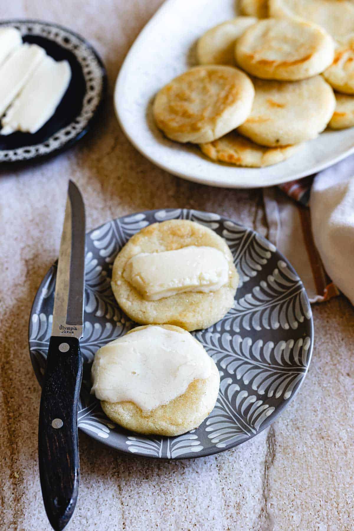 Two small round Colombian Arepas sit on a patterned plate, each topped with a slice of white cheese. A knife rests alongside them, while more arepas and cheese can be seen in the background on separate dishes.