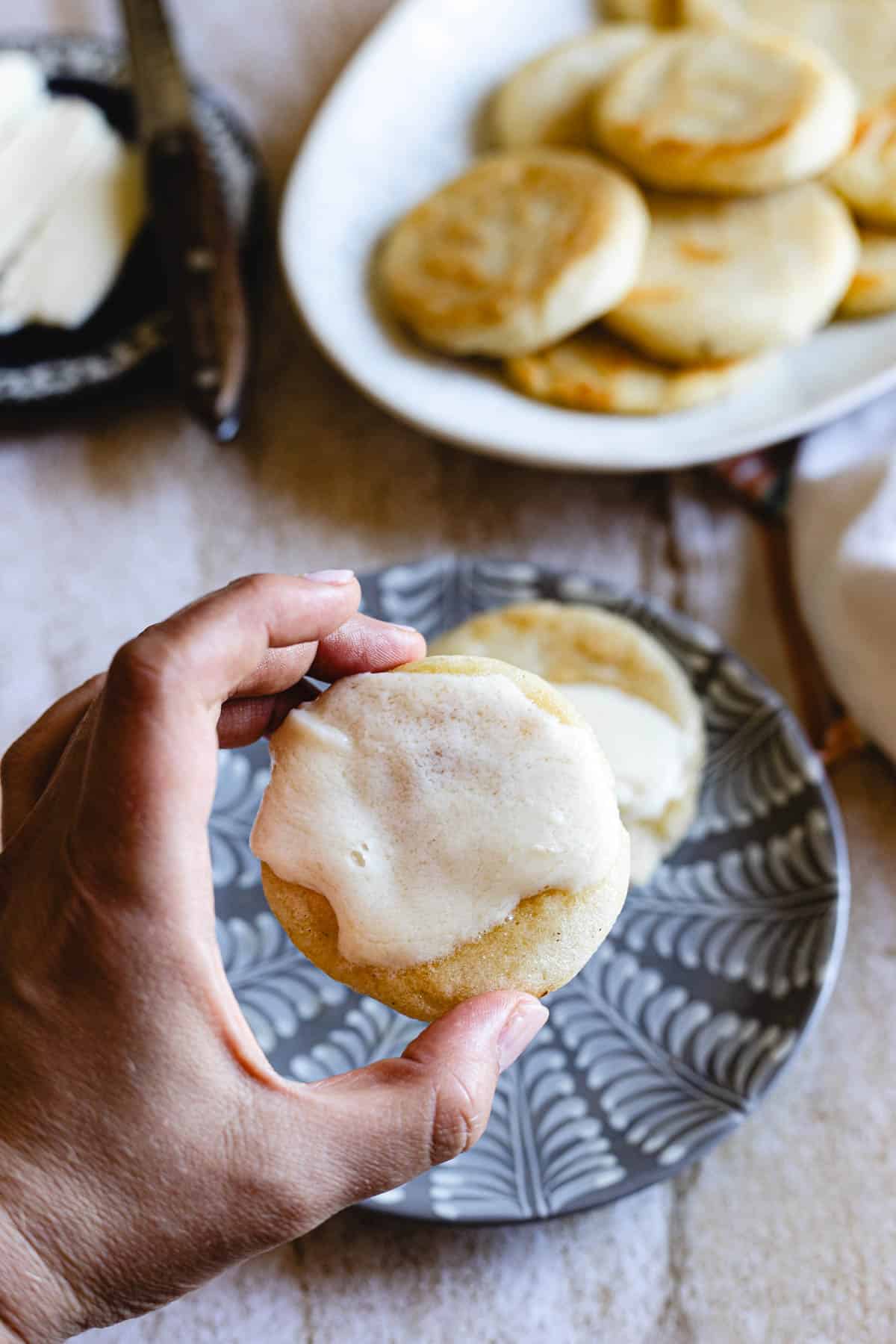 A hand holds a round Colombian Arepa topped with melted butter. More arepas are visible on a patterned plate and a white serving dish in the background. A butter knife and dish are partially visible to the side.