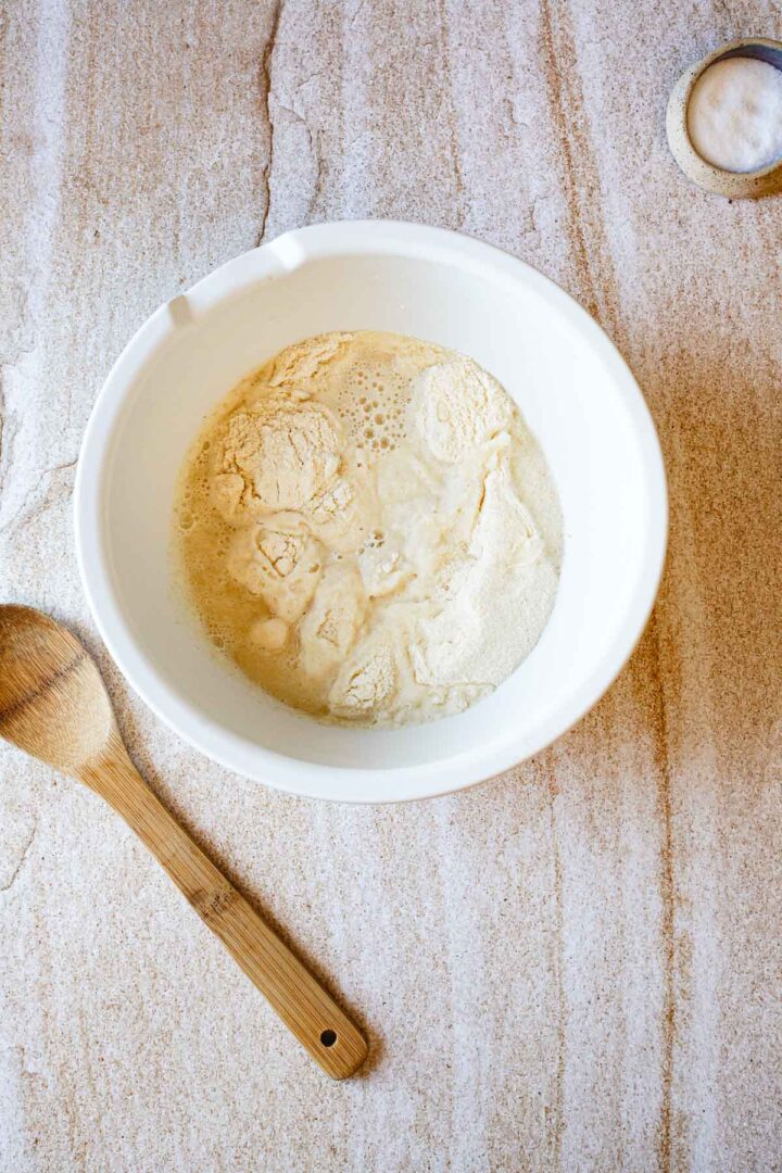 A white mixing bowl containing flour and yeast sits on a light-colored countertop, ready for making Colombian Arepas, next to a wooden spoon and a metal measuring cup filled with more flour.