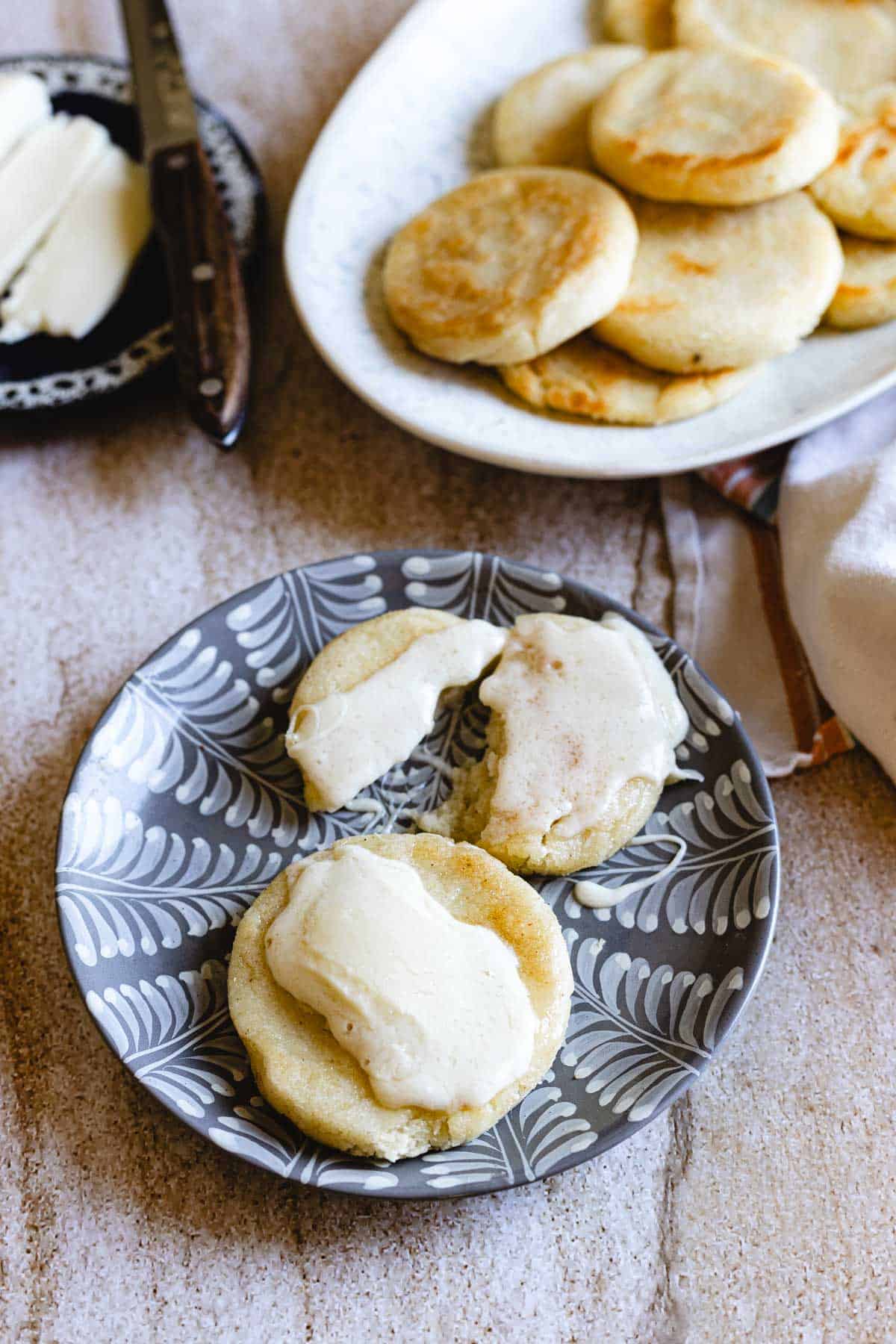 Three small round Colombian Arepas with butter rest on a patterned plate, while a larger dish of plain arepas and a butter knife with butter sit on a small plate in the background.