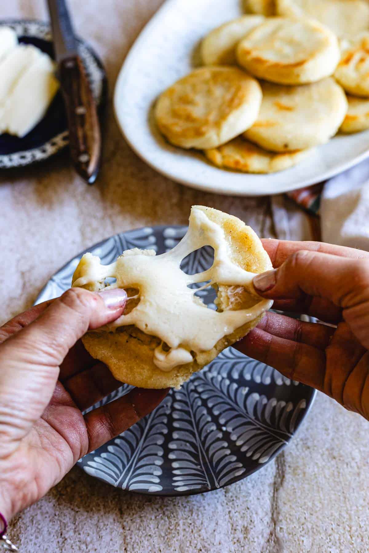 A person pulls apart a small, round Colombian Arepa, revealing melted cheese inside. A plate with more arepas and a butter dish are visible in the background.