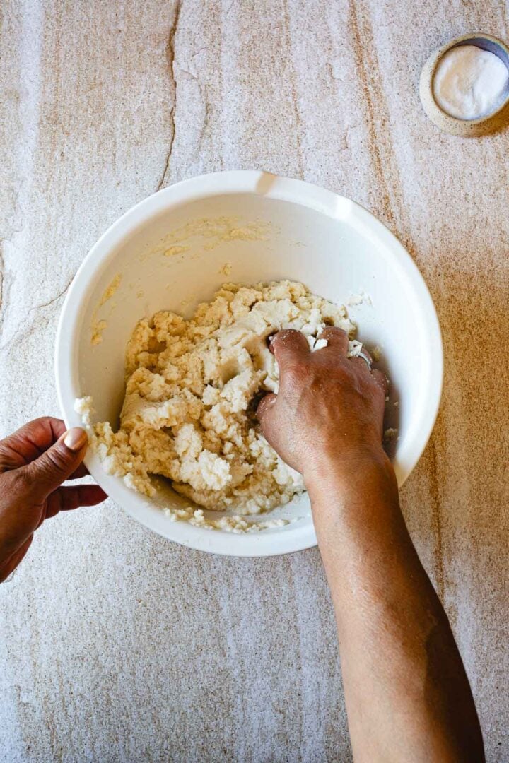 A person is mixing dough by hand in a white bowl on a light surface. A small container of salt sits in the upper right corner, illustrating a classic step in preparing a traditional Colombian food like an arepas recipe.