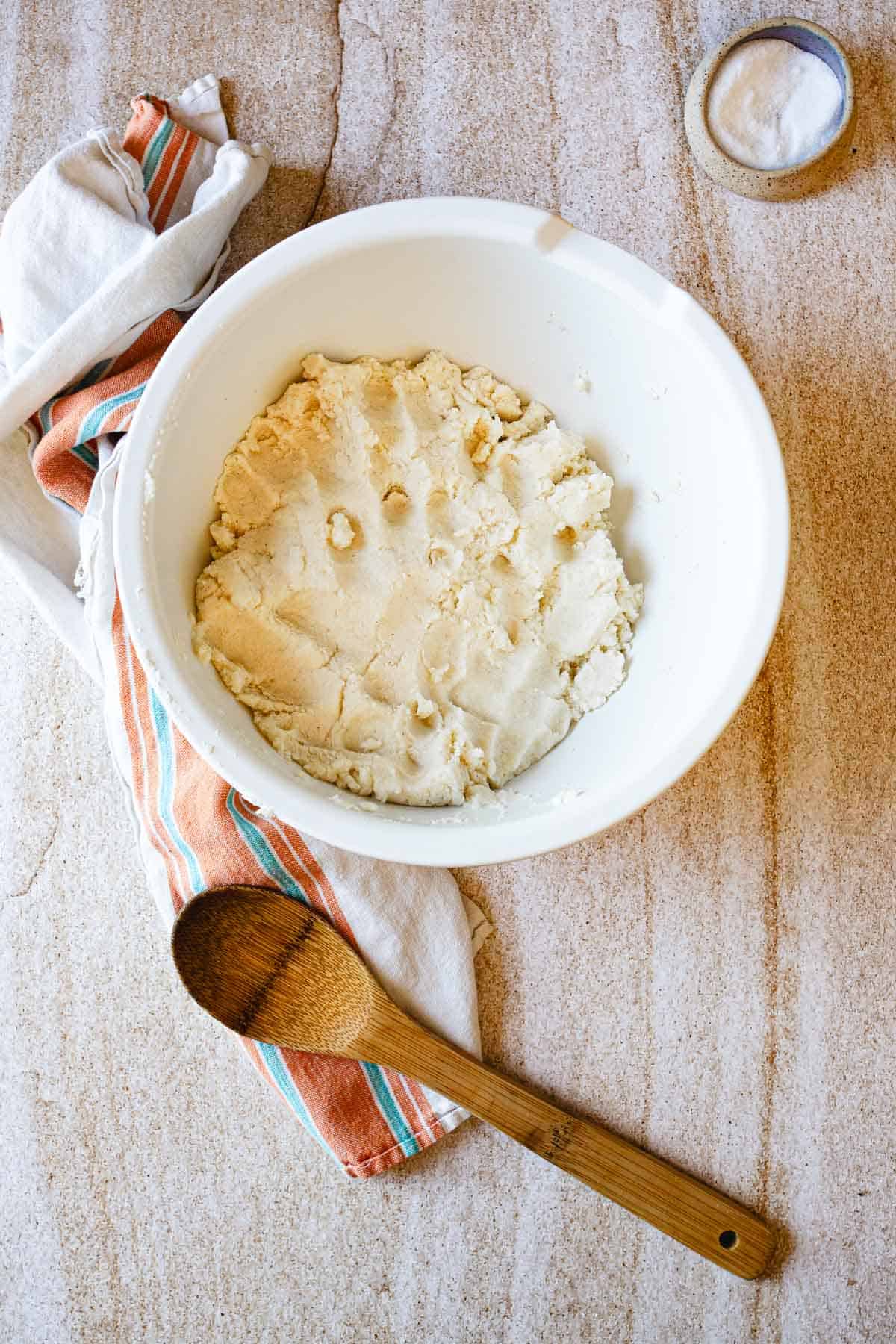 A white bowl filled with dough for Colombian Arepas sits on a countertop next to a striped dish towel, a wooden spoon, and a small bowl of salt.