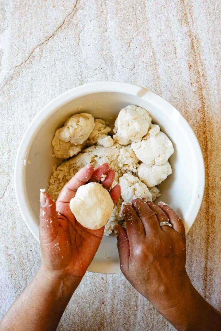 A person shapes a ball of dough with both hands over a white bowl filled with more dough on a light marble surface, preparing traditional arepas—a staple in Colombian cuisine. The dough appears slightly crumbly.
