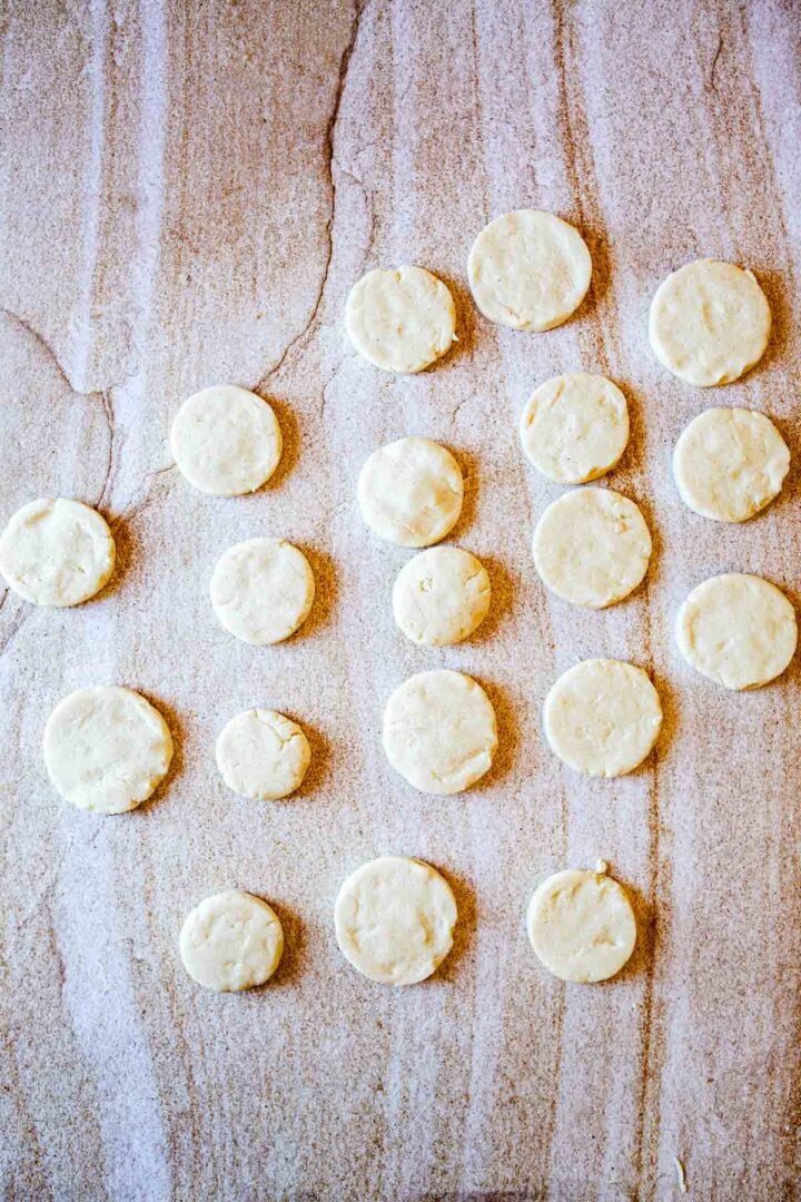 Round, unbaked biscuit dough pieces are arranged in rows on a lightly floured surface, resembling the careful preparation of a Colombian Arepas recipe. The evenly spaced dough sits atop a countertop with a subtle marble pattern.