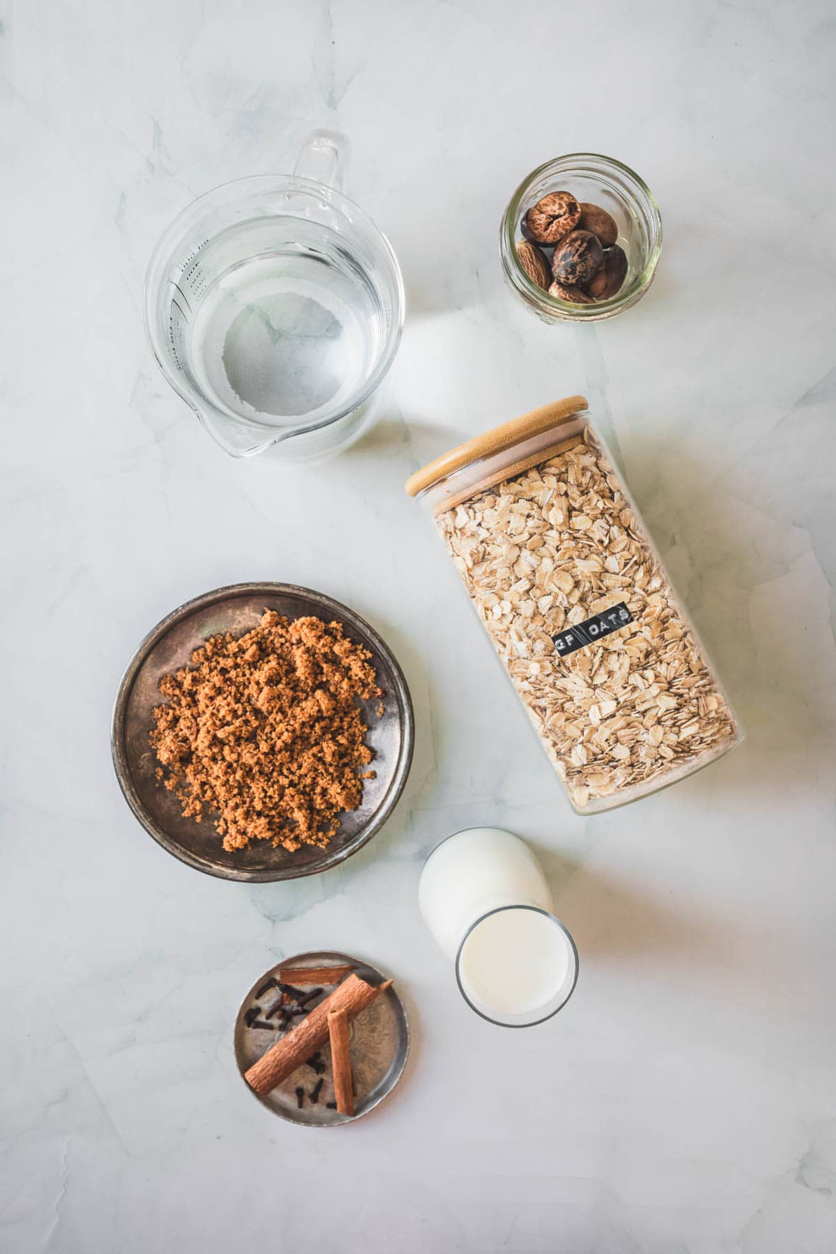 A flat lay of ingredients for Colombian oatmeal on a light surface, featuring a jar of rolled oats, a bowl of brown sugar, water, milk, whole nutmeg in a small bowl, and cinnamon sticks in a metal dish—perfect for making traditional Colombian Avena.