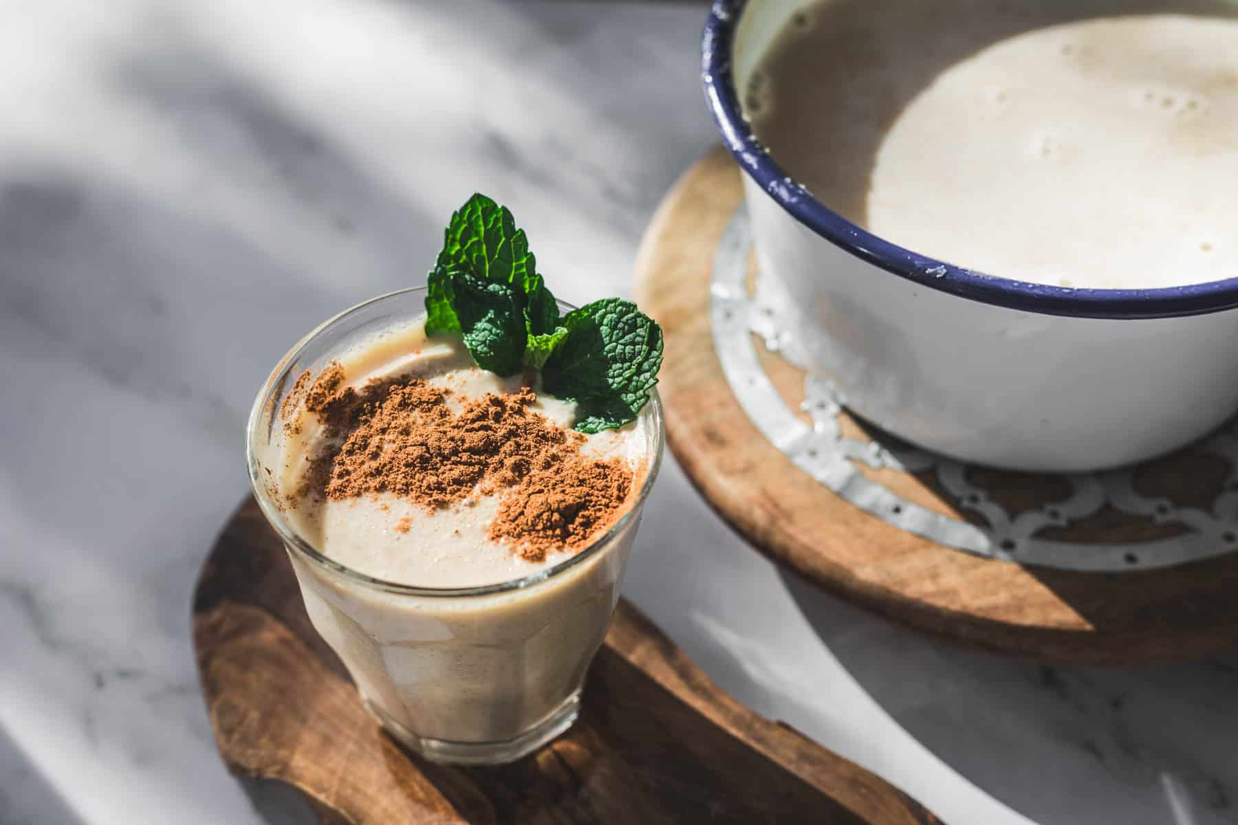 A glass of creamy Colombian Avena, a traditional Colombian drink, is topped with brown powder and fresh mint leaves on a wooden board. In the background, a white enamel pot with more Colombian oatmeal sits on a wooden trivet.