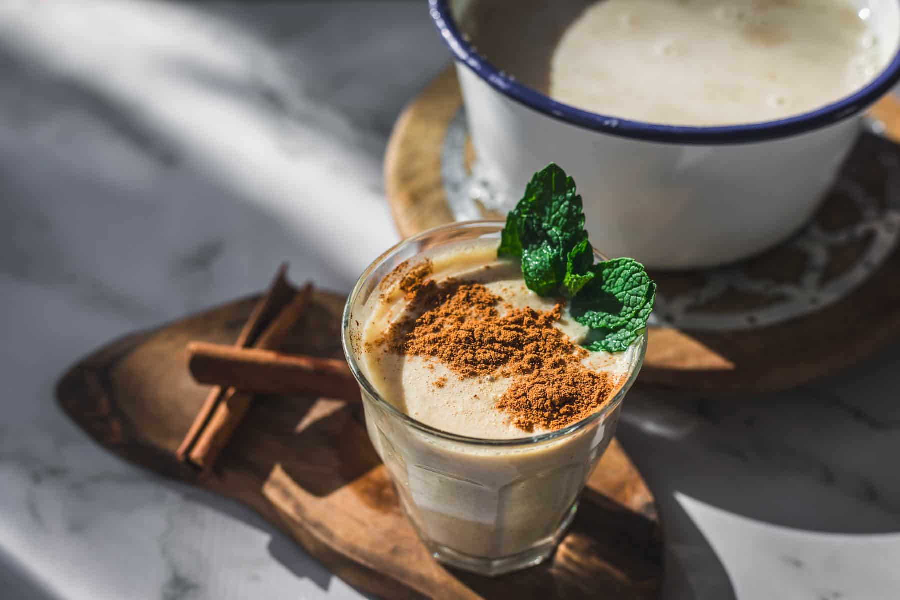 A glass of creamy Colombian Avena, a traditional Colombian drink, is topped with ground cinnamon and mint leaves on a wooden board beside cinnamon sticks, with a white enamel pot of this beloved Colombian oatmeal beverage in the background.