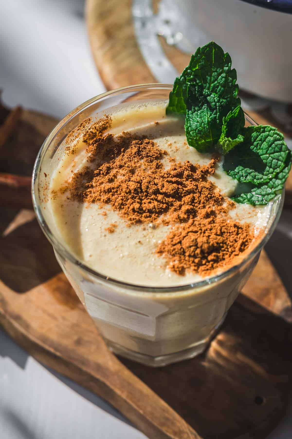 A glass filled with creamy Colombian Avena, an oatmeal drink, topped with cinnamon and garnished with fresh mint leaves, sits on a wooden board in natural light.