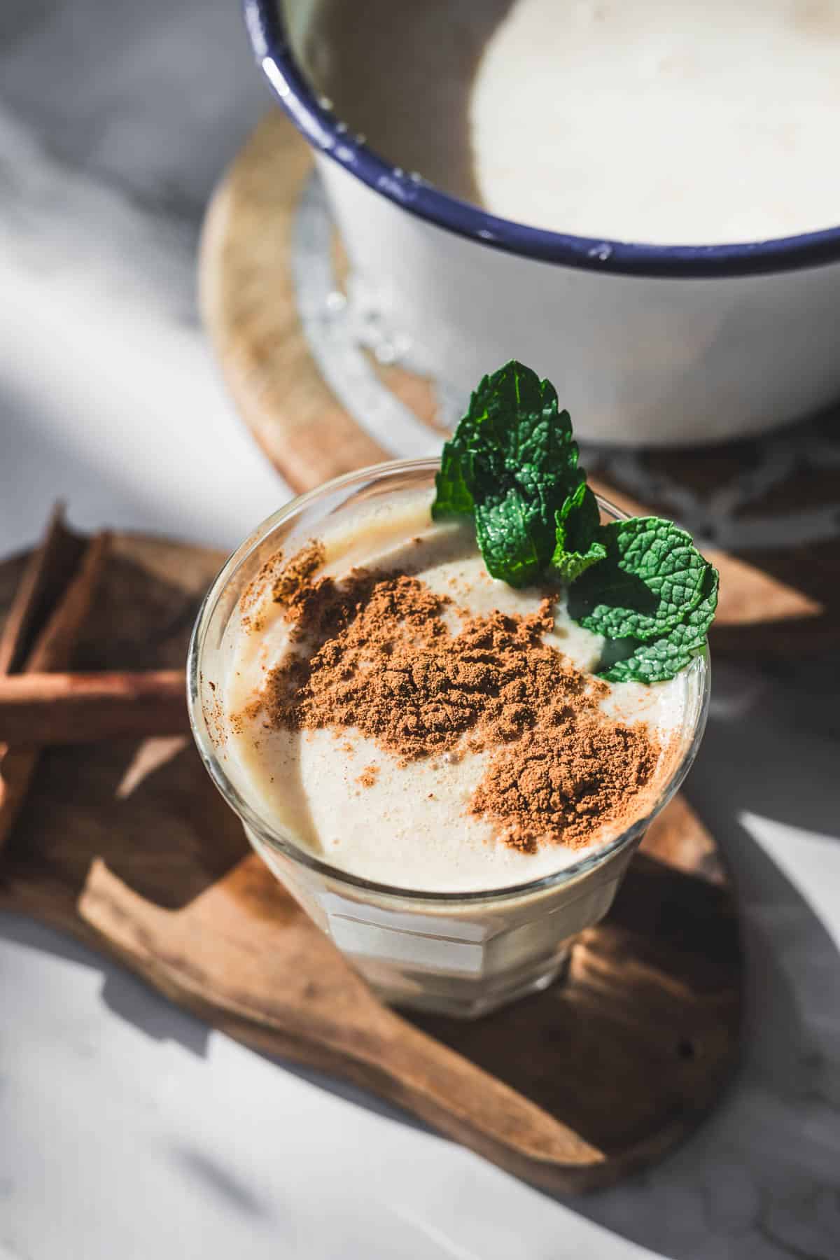 A glass of creamy Colombian Avena, a traditional Colombian drink, is topped with ground cinnamon and fresh mint leaves on a wooden board, with cinnamon sticks beside it and a pot of similar liquid in the background.