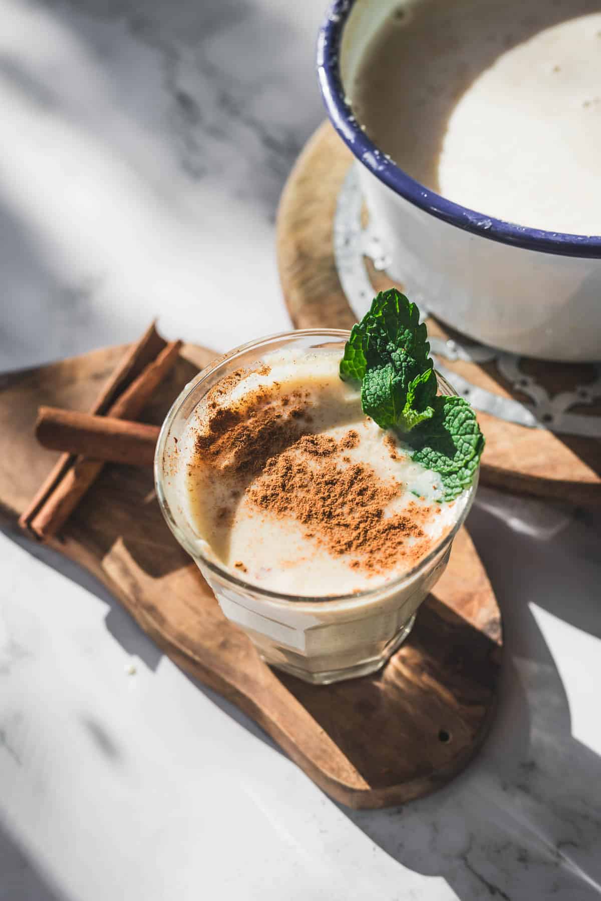 A glass of creamy Colombian Avena topped with ground cinnamon and a sprig of mint sits on a wooden board next to two cinnamon sticks, with a pot of the same traditional Colombian oat drink in the background.