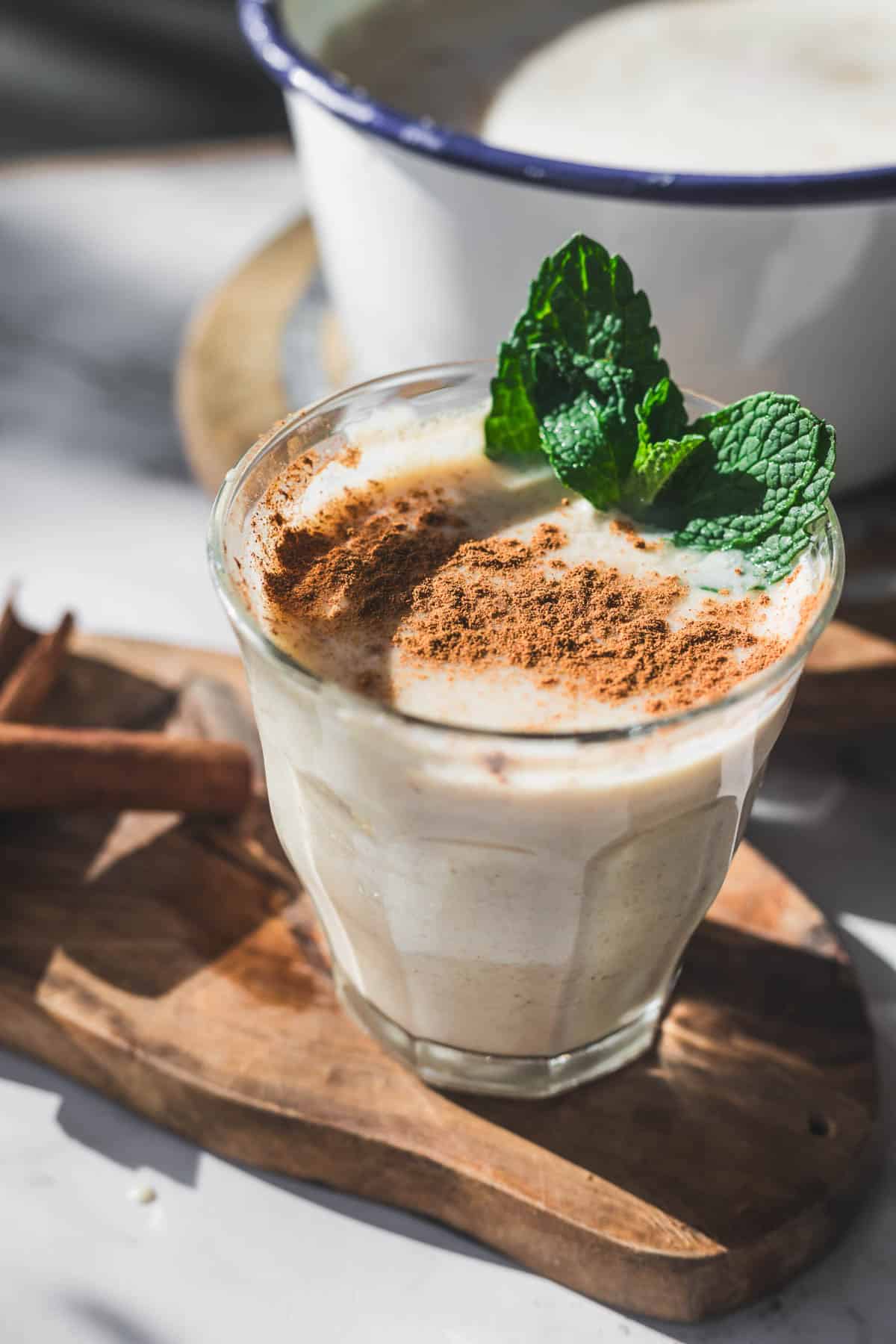 A glass of creamy Colombian Avena, a traditional Colombian drink, topped with cinnamon and a fresh mint sprig sits on a wooden board beside a cinnamon stick. A blurred pot is visible in the background.