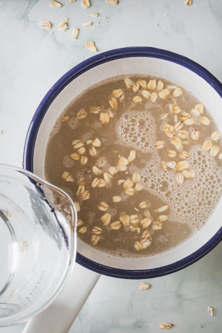 A white pot with a blue rim contains oats and water, ready to make a delicious Colombian Avena oatmeal beverage. A clear glass measuring cup sits to the left, with some oats spilled on the light-colored countertop.