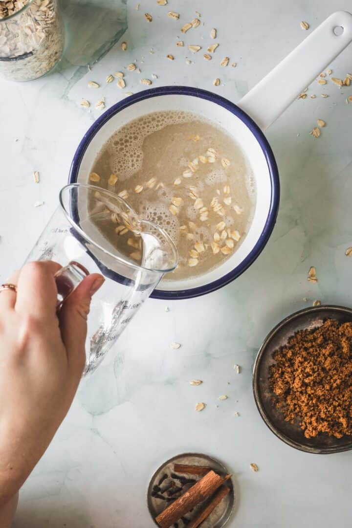A hand pours liquid from a glass measuring cup into a saucepan with oats and water, preparing Colombian Avena, a traditional Colombian drink. Brown sugar, cinnamon sticks, and more oats are arranged in small bowls on a light-colored surface.