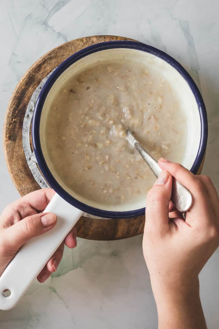 A person stirs a pot of thick, creamy Colombian Avena with a spoon. The white pot with a blue rim rests on a round wooden board atop a light marble surface.