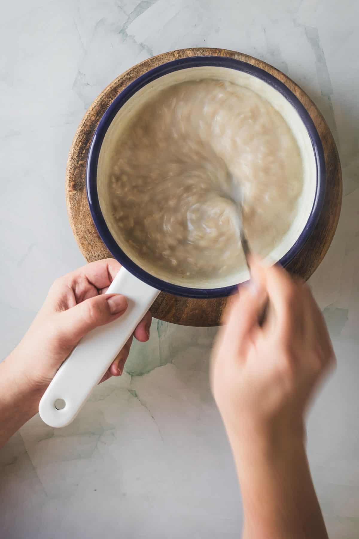 A person stirs a pot of Colombian oatmeal beverage with a spoon on a wooden board, viewed from above. The mixture appears thick and creamy, with visible motion from the stirring.