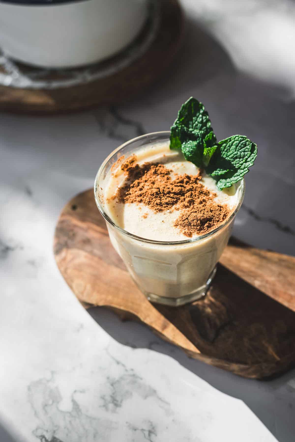 A glass of creamy Colombian Avena, a traditional Colombian drink, is topped with brown powder and two fresh mint leaves. It sits on a wooden board atop a marble surface as sunlight casts gentle shadows across the table.