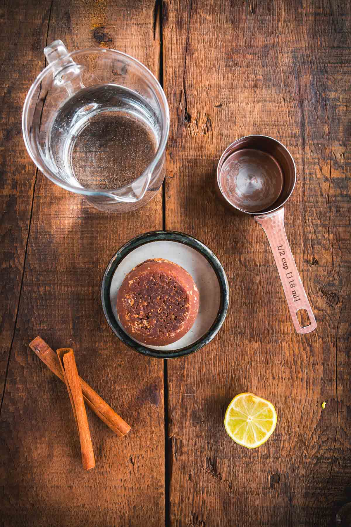 A glass pitcher of water, a metal measuring cup labeled “½ cup,” two cinnamon sticks, half a lime, and a round block of brown piloncillo on a small plate are arranged on a rustic wooden surface, perfect ingredients for an authentic Colombian Canelazo Cocktail.