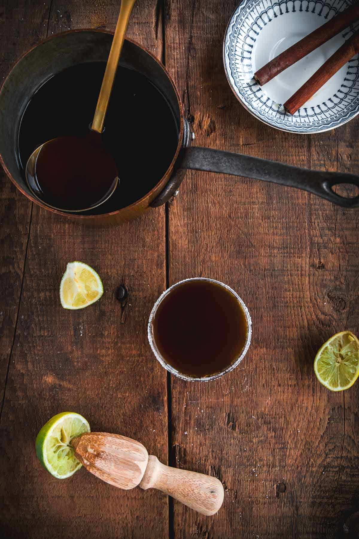 A top-down view of a wooden table with a pot of dark liquid and a ladle, a glass with the same drink, a bowl of cinnamon sticks, squeezed lime halves, and a wooden citrus reamer—perfect for preparing an authentic Colombian Canelazo Cocktail.